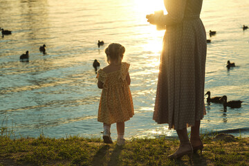 A child and woman by the lake at sunset, ducks swimming peacefully. Reflects family moments and nature's calming influence.