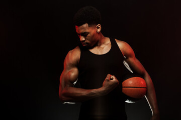 Basketball player side lit with red color holding a ball against black background. Serious muscular...