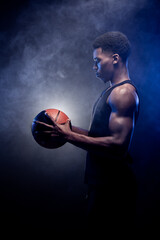 Basketball player holding a ball against blue fog background. Muscular african american man looking at basket.