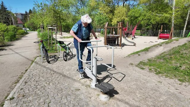 Old gray-headed senior 70-80 exercising in an outdoor creaking stepper machine in an open public garden gym park with a playground in Krakow, Poland. 4K video recorded with sound of the equipment