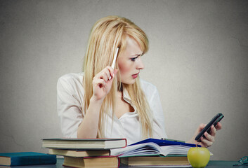 Serious young woman sitting at desk with a pile of books , reading a message on her cellphone - 784489546
