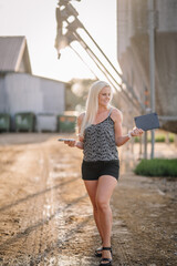 Valmiera, Latvia - August 17, 2024 - A smiling blonde woman holds a book and smartphone while standing on a farm road, with silos in the background. © Raivo