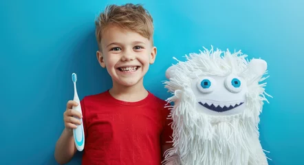 Fotobehang Cute smiling boy holding toothbrush and cute white knitted tooth pattern with eyes isolated on blue background, concept of children's dental health care © Kien