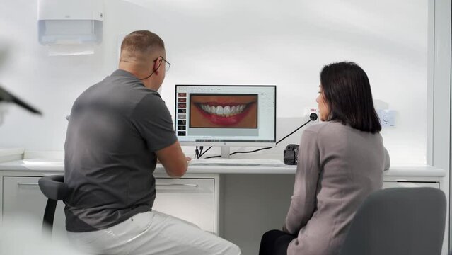 Cosmetic dentist showing photo of teeth smile to female patient on screen in modern dental clinic. Stomatologist and patient discuss modeling of veneers. Doctor explains photo of teeth to a woman.