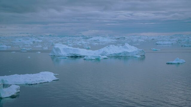 Zeitraffer von schwimmende Eisschollen bei Ilulissat in Gr&ouml;nland, ein gro&szlig;es Boot f&auml;hrt im Vordergrund vorbei, die dunklen Wolken lichten sich und die Sonne taucht das Meer in helles Licht