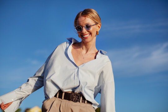 Contemporary happy woman in fancy clothes oversized shirt and sunglasses, joyfully smiling against blue sky. Trendy, happy, and stylish summer vibes