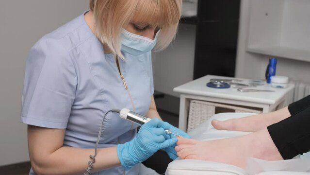 A pedicure expert in rubber gloves, performs a hardware pedicure for woman, utilizing a nail drill.
