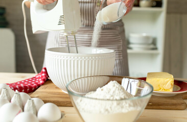 Whisking eggs with a hand mixer and pouring sugar into a bowl by a woman