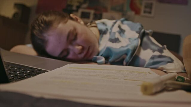 Caucasian female student fallen asleep with her head beside worksheets and laptop on bed in dimly lit bedroom at night tired of studying