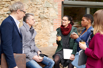 Group of multiracial colleagues talking outside cafe. Multiracial business team with drinks to go and documents communicating with each other during meeting outside cafe building
