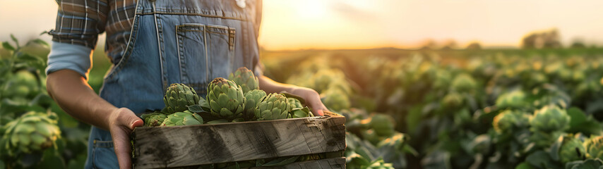 Beautiful young farmer woman holding a wooden box full of artichoke vegetable standing in the field with sunset. Concept of healthy lifestyle, local farming and beauty.