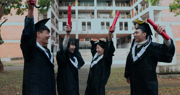 Four Asian graduates joyfully wave their diploma tubes, celebrating the completion of their studies.