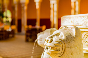 Closeup of marble head of a lion in replica of Alhambra fountain, tables and pillars in blurred...
