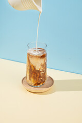 Iced coffee in a tall glass with a milk jug pouring milk on a blue and yellow background with shadow.