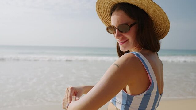 Happy young woman applying spf sun lotion on hand at seaside. Beautiful girl in straw hat putting waterproof sunscreen on skin to protect from uv. Portrait of lady in bikini using sun cream at sea.