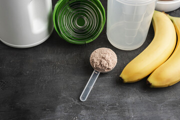 Whey protein powder with a chocolate flavor in a plastic measuring spoon on a dark background with bananas and a shaker