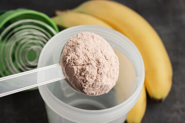 Plastic measuring spoon puts whey protein powder into a shaker on a dark background with bananas, process of making healthy protein drink