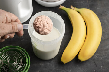 Male hand with a plastic measuring spoon puts whey protein powder into a shaker with milk on a dark...