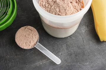 Whey protein powder with a chocolate flavor in a plastic measuring spoon, shaker with a milk and bananas on a dark background, process of making protein shake drink