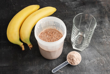 Blended chocolate protein drink in a shaker, plastic measuring spoon with protein powder, bananas and drinking glass on a dark background