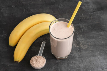 Blended chocolate protein drink in a glass with yellow straw, plastic measuring spoon with protein powder and bananas on a dark background