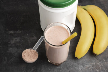 Blended chocolate protein drink in a glass with yellow straw, plastic measuring spoon with protein powder and bananas on a dark background