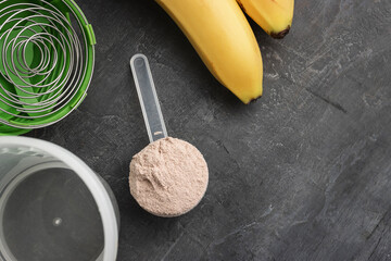 Whey protein powder with a chocolate flavor in a plastic measuring spoon on a dark background with bananas and a shaker, top view