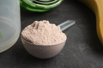 Whey protein powder with a chocolate flavor in a plastic measuring spoon on a dark background with bananas and a shaker