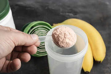 Male hand with a plastic measuring spoon puts whey protein powder into a shaker on a dark background with bananas, process of making healthy protein drink