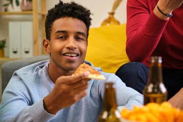 African young man eating pizza, smiling and looking at camera at shared flat.