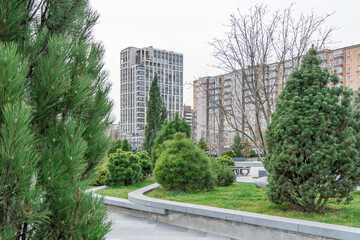 Nice modern leisure city park with benchs surrounded high-rise building. Public park in town residential. Sidewalk and walkway with conifer trees. Place to rest in city landscape. Urban architecture.