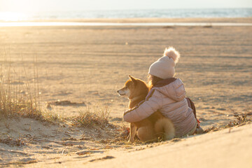 A young girl with a dog in nature. Kid girl sitting with a shiba inu dog on the beach at sunset in Greece in winter - 784626755