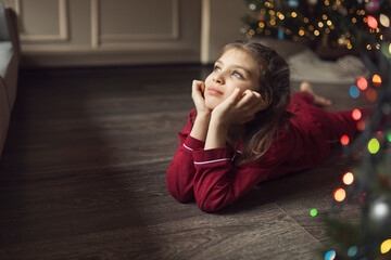 Girl of 7 years old in red pyjamas lies on floor of house and looks out of window next to Christmas...
