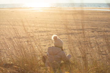A child girl wearing the hat with a pompon sitting on the beach and looking at the sea in sunset time back to the camera - 784630312