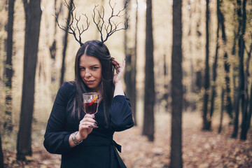 Mysterious woman wearing gothic deer antlers headband with mulled wine standing in autumn forest