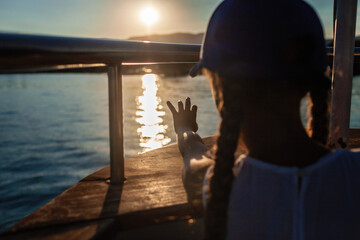 Child in a blue cap at sunset on a boat, touching sunlight reflection on the water, evoking a sense...