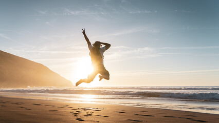 Happy man with backpack jumping with arms raised on the beach at sunset - Charming tourist enjoying...