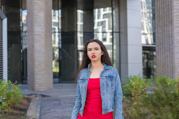 A young girl in a red dress stands in front of a building. She's wearing a denim jacket and red lipstick.