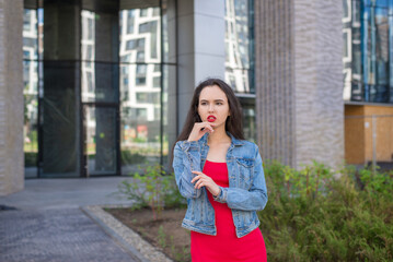 Fototapeta premium A young girl in a red dress stands in front of a building. She's wearing a denim jacket and red lipstick.