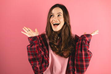Optimistic woman raises palms from joy, happy to receive awesome present from someone, shouts loudly, dressed in plaid shirt, isolated on pink background. Excited female yells, raised hand, look side.