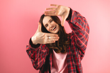 Attractive woman with a lovely smile making a frame gesture with her fingers framing her face. Woman posing on selfie photo looking at camera standing isolated pink background. Girl wear plaid shirt.