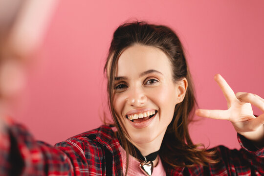 Close up photo of beautiful young brunette woman with high ponytail taking selfie, posing with peace v-sign, smiling happy, take photo, straight hand to camera posing against pink background.