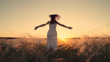 Free girl runs happily through meadow in grass in rays of sunset. Concept of female dreams, success, travel, flight. Young woman holding her arms to sides runs across field in sun. Happy running girl