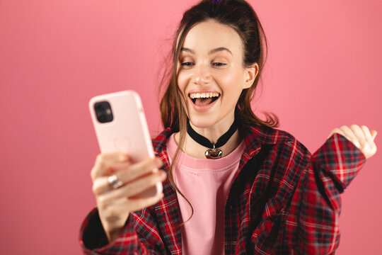 Amazed brunette woman with high ponytail look surprised wow on pink background. Girl look on mobile phone and doing winner gesture. Girl just found out big win news, win gesture.