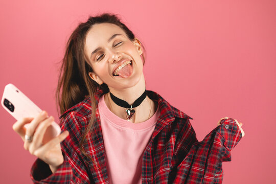 Amazed brunette woman with high ponytail look surprised wow on pink background. Girl look on mobile phone and doing win gesture. Girl just found out big win news, win gesture, tongue out, closed eyes.