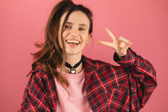 Smiling woman make peace sign against pink background. Brunette woman with high ponytail wear pink long sleeves and red plaid shirt. Happy laughing girl show v victory gestures, dancing, have fun.
