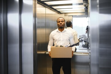 A young african man fired from work is standing in the elevator with a box of things