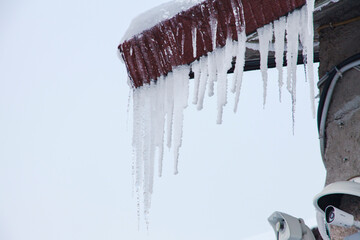 Long icicle formations hanging from the eaves of a building’s roof, with security cameras below.

