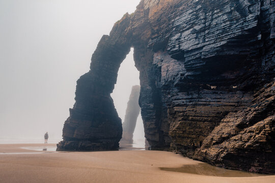 Tourist walking along natural arch on Cathedrals beach in Galicia, Spainn. Man silhouette in foggy landscape with Playa de Las Catedrales Catedrais beach in Ribadeo, Lugo on Cantabrian coast