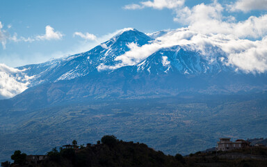 Vue sur l'Etna sous le soleil avec quelques nuages, et des maisons sur une colline au premier plan